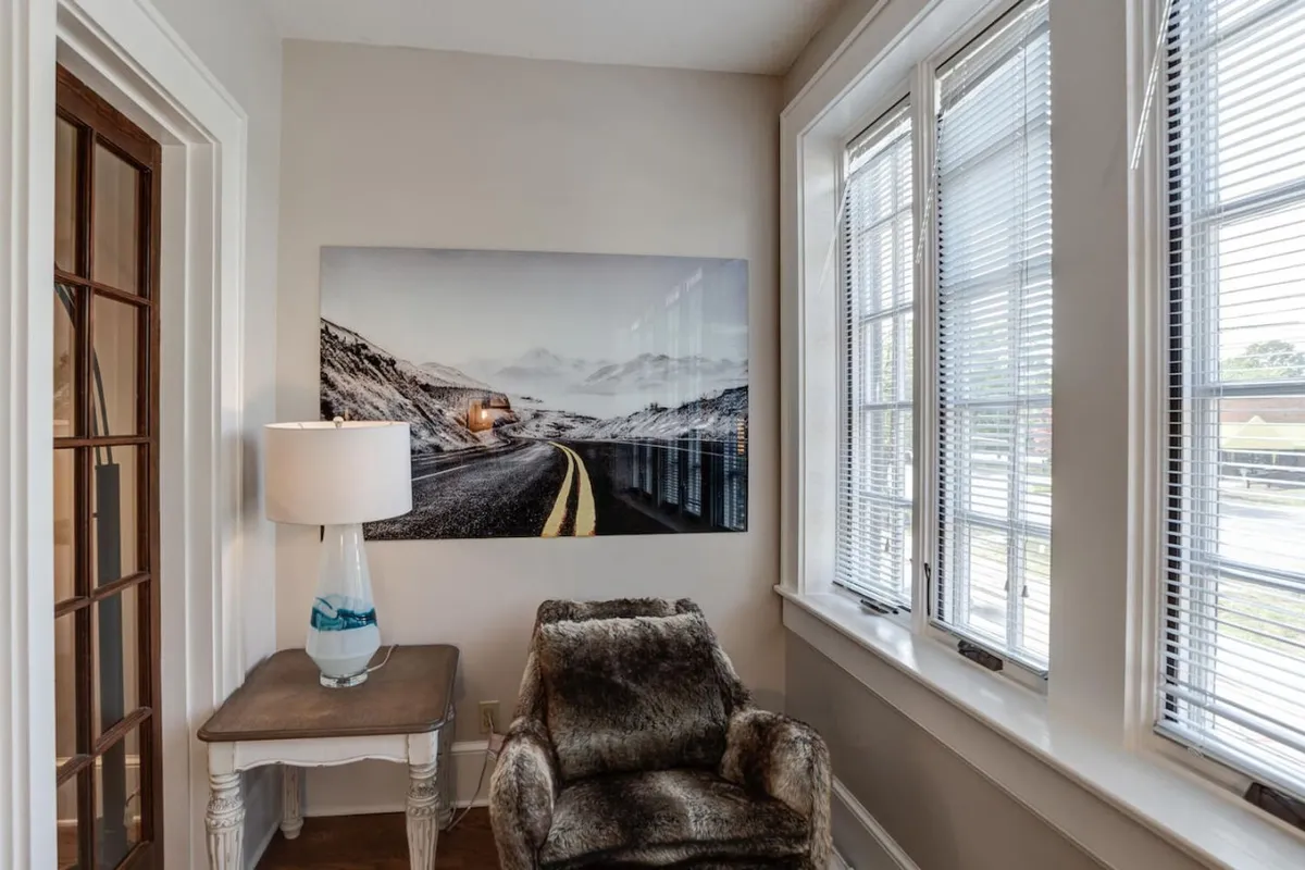 This image presents a cozy reading nook with a comfortable chair, table, and a lamp, set beside a window. The focal point is an artwork depicting a winding road, subtly alluding to exploration and the journey beyond the condo. Natural light pours in, inviting both relaxation and daydreaming about the city just steps away.