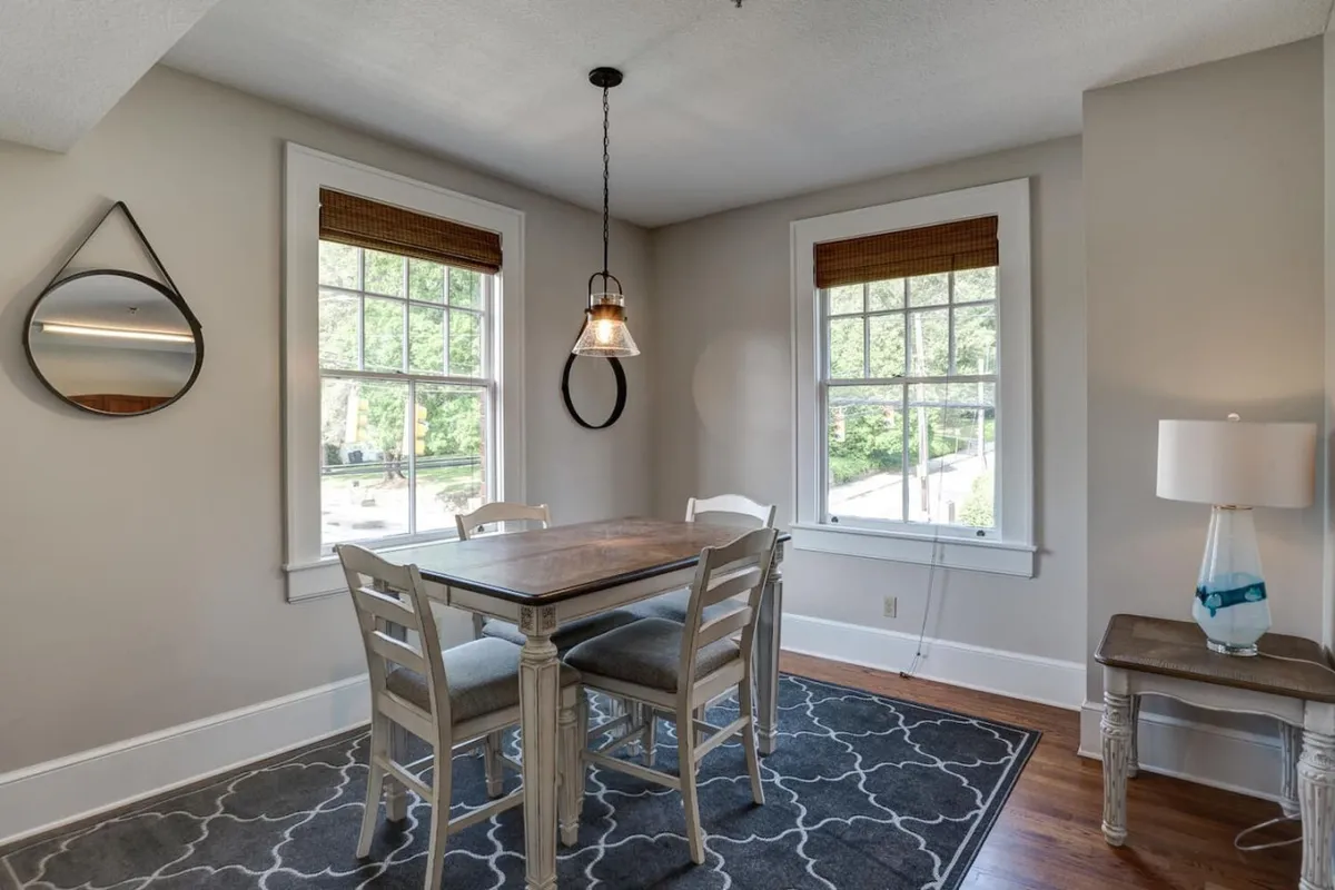 A cozy and welcoming dining area featuring a large table surrounded by chairs, with expansive windows that flood the space with natural light. Stylish pendant lighting hangs above, and a mirror and lamp add warmth and ambiance. The setting suggests a versatile space perfect for casual meals, working, or socializing with friends.