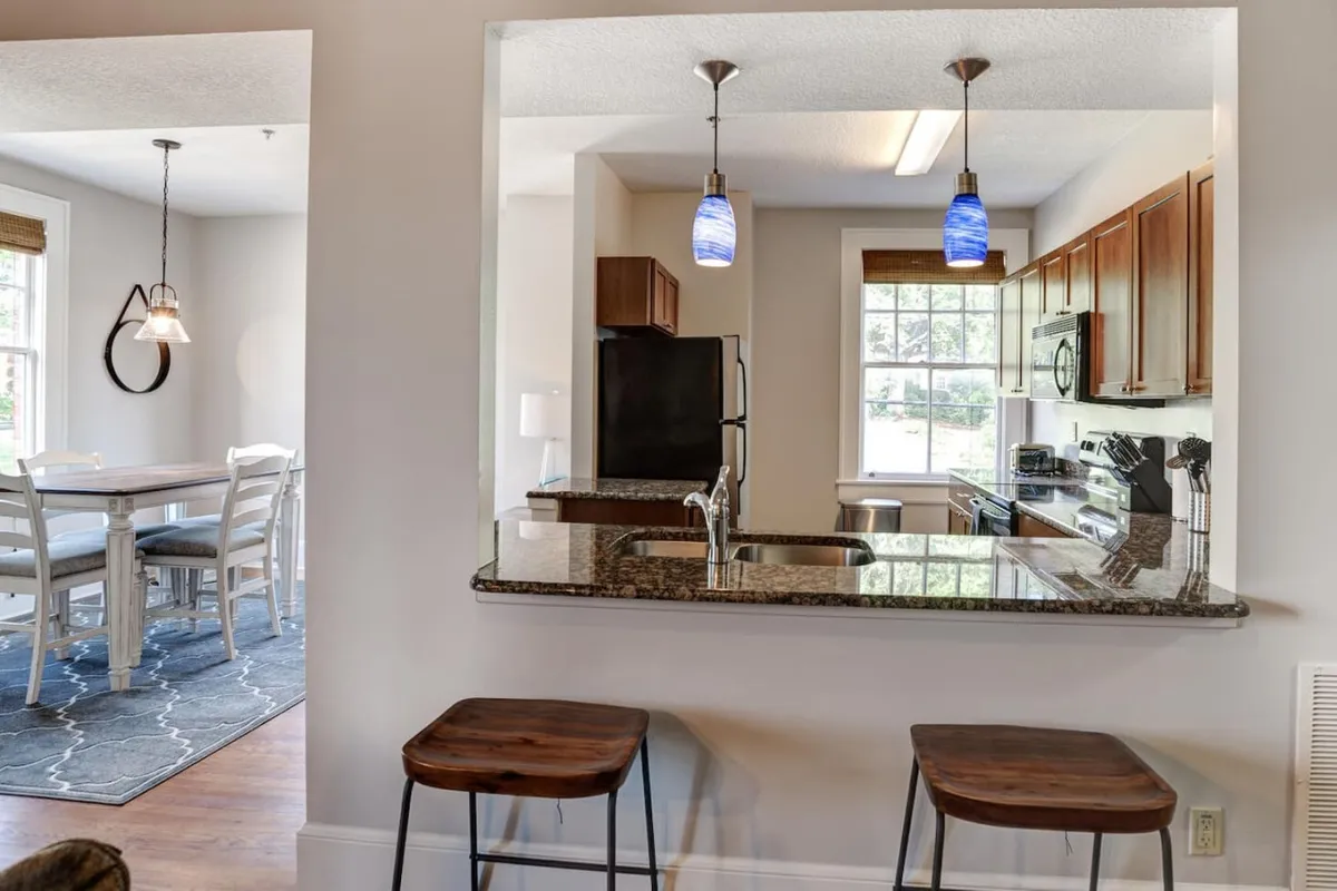 A modern kitchen scene with a spacious island bar featuring stools, high-end appliances, and a glimpse of the adjacent dining area. The layout encourages casual dining and social interaction, suggesting a lively atmosphere where guests can gather, cook together, or unwind after dining out.