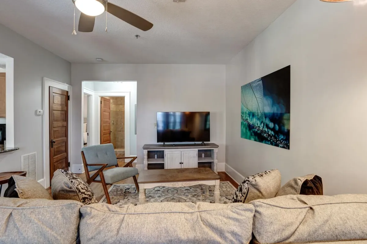 The image captures a stylish living area with a plush sofa, modern coffee table, large television, and vibrant artwork on the wall, all set in a cozy, well-lit space perfect for unwinding. The room exudes warmth and character, inviting guests to relax and enjoy intimate moments or movie nights after a day exploring Asheville.