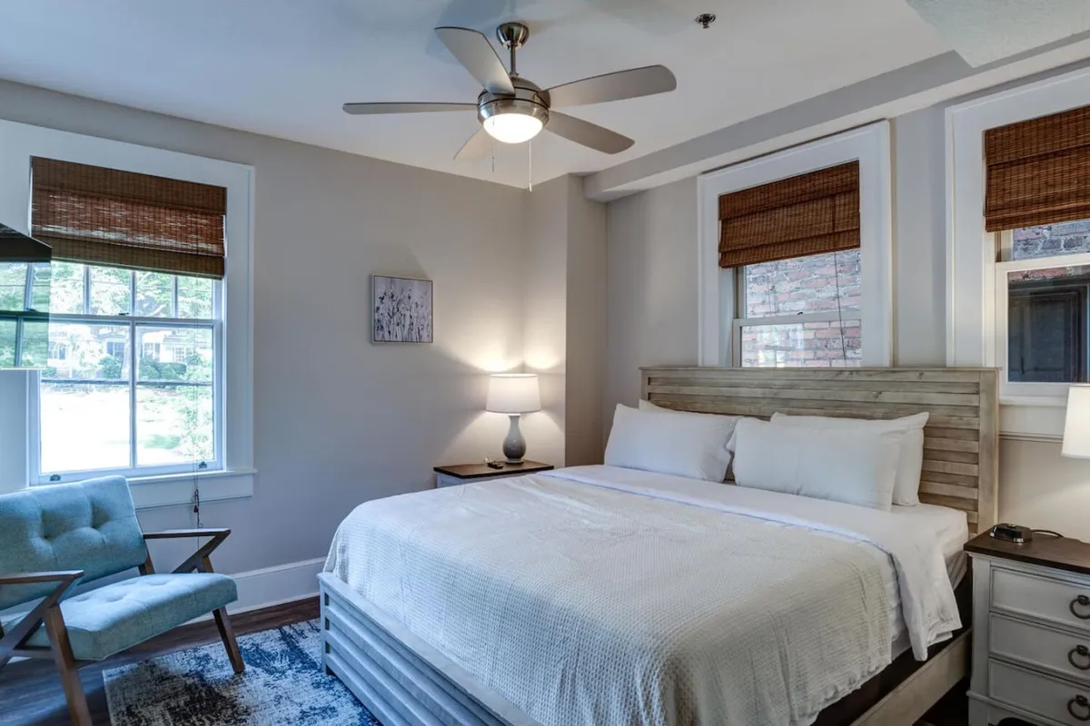 A tranquil bedroom featuring a comfortable bed dressed in crisp white linens, a wooden headboard, a blue accent chair beside the bed, and a nightstand with a lamp. Soft natural light filters into the room, highlighting the calming, restful atmosphere.