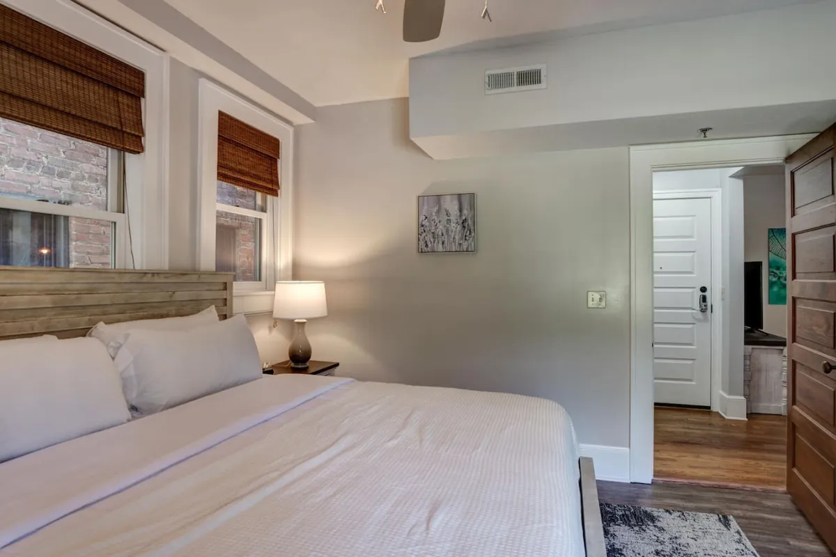 A peaceful bedroom view with a plush bed, bedside lamp, tasteful artwork, and double doors. Natural light brightens the space, enhancing the sense of tranquility and comfort.