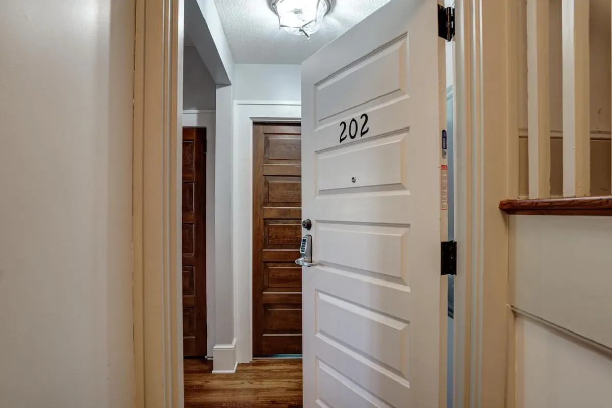 A view of an open apartment door with the number 202, flanked by warm wooden doors and illuminated by a soft overhead light. The entryway is bright and welcoming, hinting at the transition from the bustling city outside into a private residence.