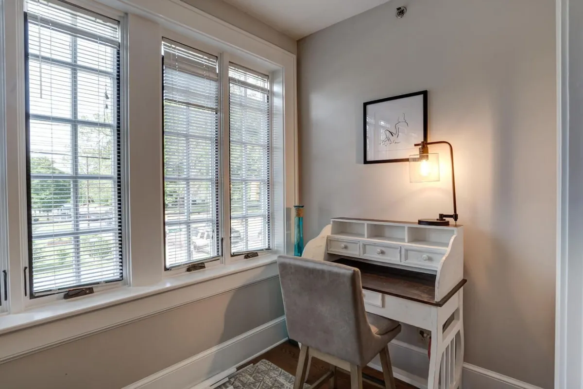 The photo shows a bright workspace with a desk set against large windows, letting in abundant daylight. Stylish artwork and a modern light fixture add to the serene and productive atmosphere, suggesting a place to recharge and plan your next outing.